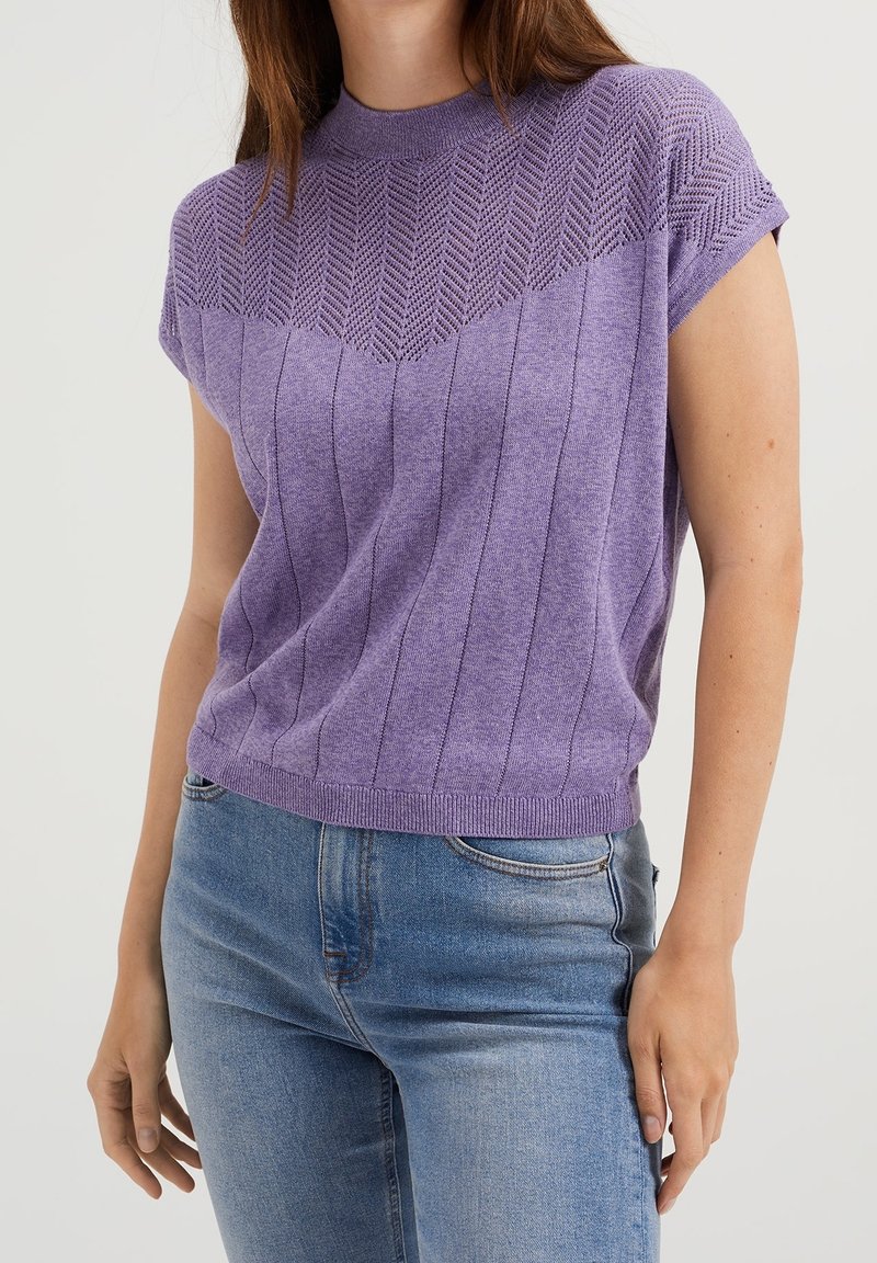 Kvinna iklädd en kortärmad lila stickad topp med sicksack- och vertikala randmönster, tillsammans med ljusblå denimjeans.