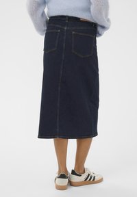 Dunkelblauer Denimrock in Midi-Länge mit zwei Gesäßtaschen und gelben Nähten. Kombiniert mit weißen Schuhen mit schwarzen Akzenten.