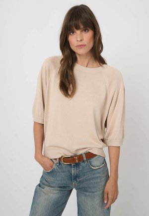 Kobieta o długich brązowych włosach, ubrana w beżowy sweter z krótkim rękawem i niebieskie dżinsy z brązowym paskiem, pozująca na tle jednolitego jasnego tła.