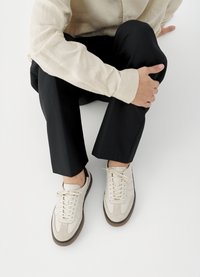 Baskets en daim et cuir beige avec des lacets blancs, associées à un pantalon noir sur mesure et une chemise boutonnée de couleur claire.
