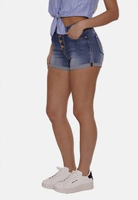 Shorts in denim di blu medio con un design aderente, orli arrotolati e cinque bottoni in vita. Abbinati a sneakers bianche.