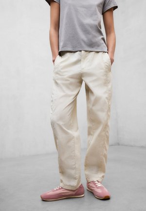 Personne portant un pantalon ample beige clair, un t-shirt gris à manches courtes et des baskets roses, debout les mains dans les poches sur un fond gris.