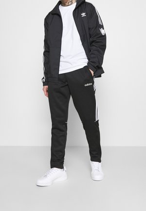 Mann trägt eine schwarze Adidas Trainingsjacke und -hose mit weißen Streifen, kombiniert mit weißen Sneakers und einem weißen T-Shirt.