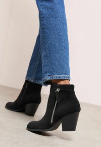 Botas de tobillo de ante negras con un tacón grueso, que cuentan con una cremallera lateral y un acabado texturizado. Combinadas con unos pantalones vaqueros de mezclilla azul claro.