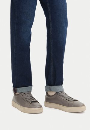 Szare skórzane trampki o gładkiej fakturze, z okrągłym noskiem i białą gumową podeszwą; noszone z ciemnymi dżinsami z mankietami przy kostkach.