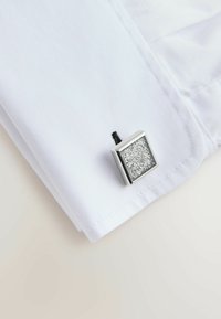 Polsino di una camicia bianca con un gemello quadrato argento che presenta una superficie texture luccicante, fissato attraverso un'asola.