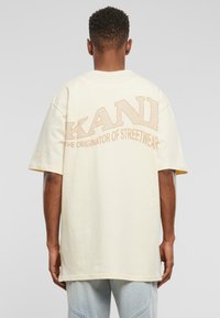 Light yellow oversized cotton t-shirt with large, textured "KANI" logo in beige on the back and "THE ORIGINATOR OF STREETWEAR" below it.