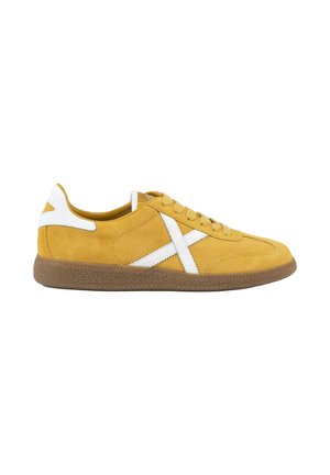 Zapatilla amarilla con suela marrón, detalle blanco en el talón y diseño en forma de X blanco cosido en el lateral, mostrada desde el lado exterior.