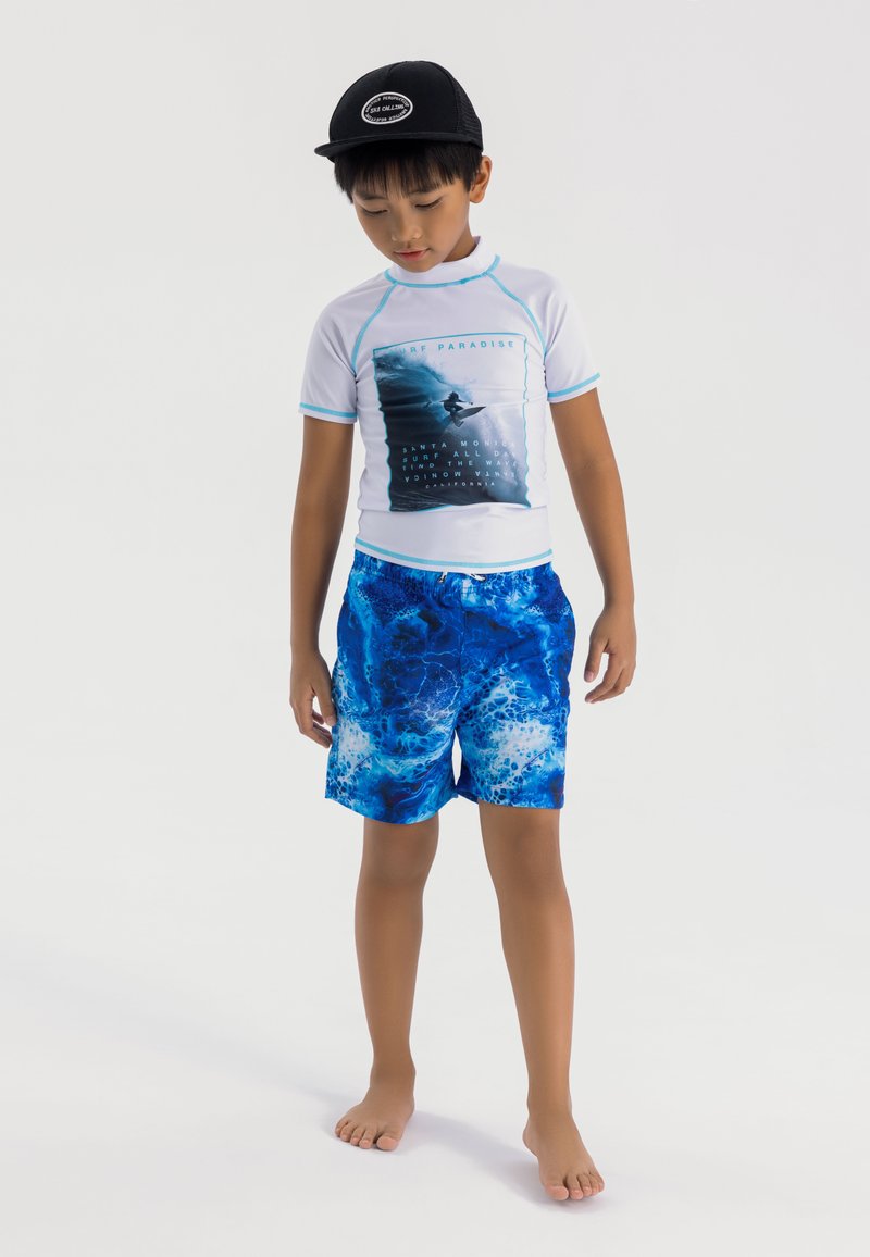 Vit rash guard med kort ärm och blåa detaljer, prydd med grafik framtill, ihop med blå badshorts med ett virvlande havsmönster. Svart keps.