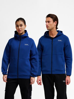 Un homme et une femme portant des sweats à capuche bleus assortis avec fermeture éclair et pantalons noirs, debout côte à côte sur un fond blanc.