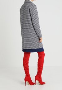 Osoba v kostkovaném černobílém kabátu přes tmavě modré šaty a červených semišových lodičkách na vysokém podpatku, stojící proti jednobarevnému pozadí.