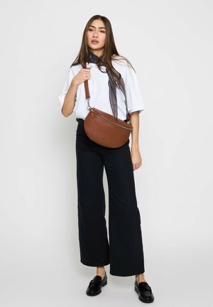 Young woman wearing white oversized T-shirt, black wide-leg pants, black loafers, patterned scarf, and holding a brown leather crossbody bag.