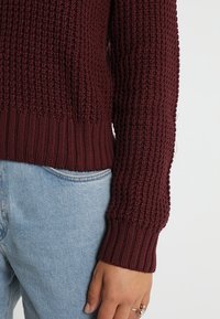 Bordowy sweter z dzianiny z teksturowanym wzorem, ściągaczami na rękawach i dopasowanym dołem, zestawiony z jasnoniebieskimi dżinsami.