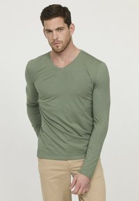 T-shirt à manches longues, vert olive, fabriqué en tissu doux. Présente un col en V discret et un petit logo sur la poitrine gauche. Design ajusté.
