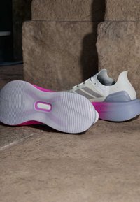 Scarpa con una parte superiore bianca e accenti grigi, una suola texturizzata specializzata con sfumature di rosa e viola, e un design del tallone curvato.