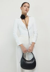 Schwarze Ledertasche mit geschwungener Silhouette, glatter Textur und kurzem Griff. Das Model trägt einen weißen knöpfbaren Body mit einem schwarzen Rosen-Choker.