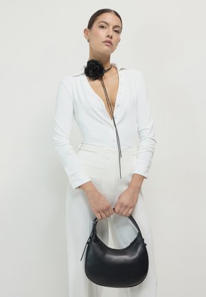 Schwarze Ledertasche mit geschwungener Silhouette, glatter Textur und kurzem Griff. Das Model trägt einen weißen knöpfbaren Body mit einem schwarzen Rosen-Choker.