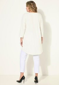 Tunic a maniche lunghe color beige chiaro con orlo arrotondato, abbinata a pantaloni bianchi fitted e scarpe con tacco alto nere. Tessuto dalla texture liscia.
