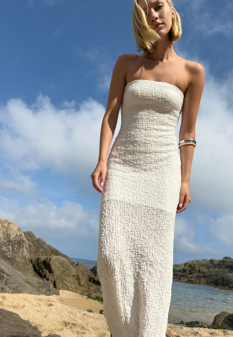 Abito bianco senza spalline con tessuto strutturato, corpino aderente e gonna fluente. La modella indossa un bracciale a manica in argento in un ambiente da spiaggia.