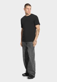 Czarna koszulka z krótkim rękawem w połączeniu z szarymi luźnymi dżinsami i czarnym obuwiem. Posiada prosty design bez widocznych logo czy wzorów.