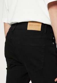 Pantalones vaqueros de mezclilla negra con una textura suave, que cuentan con una etiqueta de cuero marrón etiquetada "ONLY & SONS" en la cinturilla y dos bolsillos traseros.