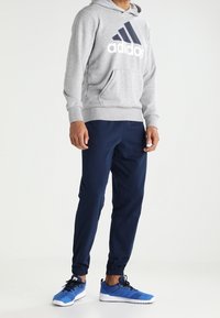 Sweat à capuche gris avec un grand logo blanc "adidas", associé à un pantalon de jogging bleu marine. Le pantalon de jogging a des chevilles élastiques, porté avec des chaussures de sport bleues.
