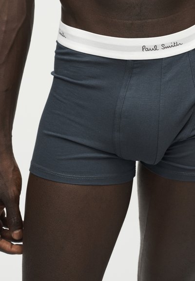 Gros plan d'une personne portant un boxer bleu foncé avec une ceinture blanche comportant le logo Paul Smith.