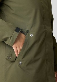 Olivengrøn jakke med en glat tekstur, der har en sidelomme sikret med en trykknap og Velcro-detaljer ved manchetterne.