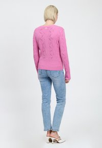 Donna con capelli biondi corti, che indossa un maglione rosa a trama, jeans azzurri chiari e mule bianche con tacco alto, in piedi e girata di spalle.