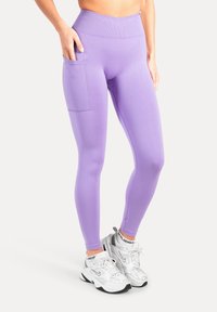 Paarse leggings met een hoge tailleband, voorzien van een zijzak en gemaakt van een gladde, rekbare stof. Gecombineerd met witte sportschoenen.