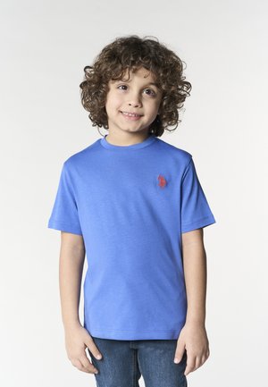 Bambino sorridente con capelli ricci che indossa una maglietta blu a maniche corte e jeans, in piedi davanti a uno sfondo chiaro e uniforme.