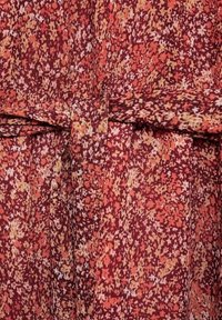 Gros plan d’un tissu avec un motif floral dense et petit dans des tons rouges, orange, beige et blanc, avec une ceinture en tissu nouée à la taille.