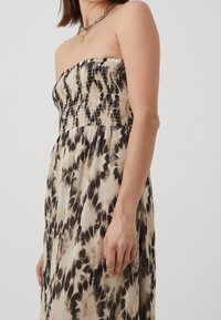 Nainen, joka on pukeutunut olkaimettomaan mekkoon, jossa on kimppu vartaloa ja beige-mustaa abstraktia eläinkuosia, asusteena kerrokselliset hopea- ja vihreät kaulaketjut.