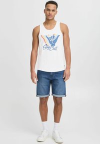 White tank top featuring "Stay Chill" graphic with multi-colored hand sign; paired with rolled denim shorts and white sneakers.