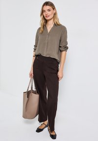 Khakifarbenes Hemd mit Knöpfen, schwarze flache Schuhe und braune weite Hosen. Beiger Tote-Bag. Glatter Stoff und lässiger Schnitt.