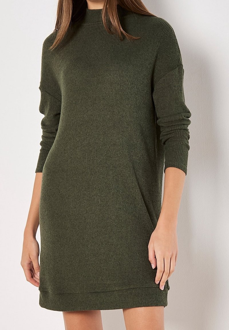 Femme portant une robe pull en maille côtelée olive verte à manches longues et col montant, debout devant un fond clair uni.