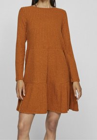 Femme portant une robe longue à manches longues, côtelée, de couleur rouille, avec une jupe à étages, debout devant un fond gris clair uni.