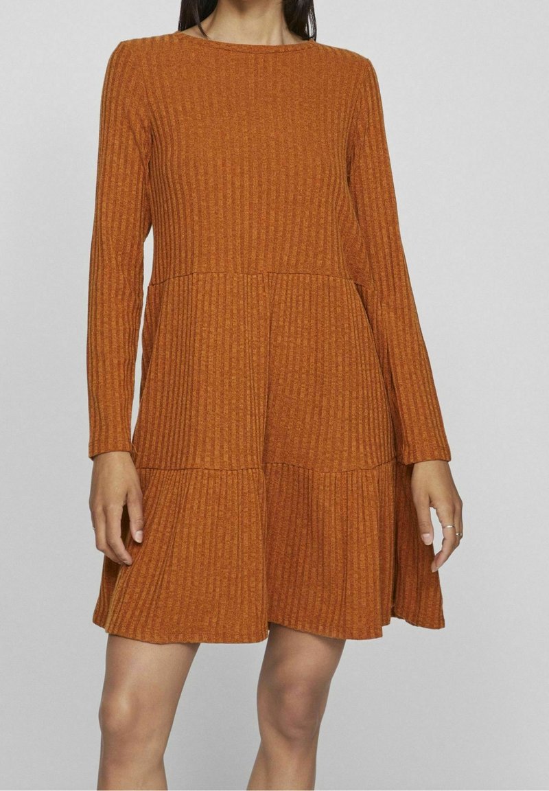 Femme portant une robe longue à manches longues, côtelée, de couleur rouille, avec une jupe à étages, debout devant un fond gris clair uni.