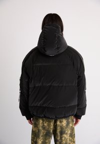 Svart dunjakke med stor hette, med quiltede paneler og en glatt tekstur. Brukt med bukser i camouflage-mønster.