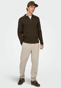 Brązowy sweter na zamek, jasnobeżowe spodnie dresowe i brązowe buty na sznurowadła. Model nosi brązową czapkę. Dzianinowa faktura i kołnierzyk.