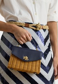 Ruka drži malu tkanu torbicu s tamnoplavim preklopom i zlatnim RLL logotipom, odjevena u suknju s morskim i bijelim chevron uzorkom i uže remenom.