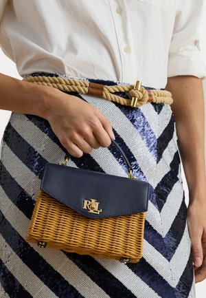 Mão segurando uma pequena bolsa de palha com aba azul-marinho e logótipo dourado RLL, vestindo uma saia com padrão em zigue-zague azul-marinho e branca, adornada com um cinto de corda.