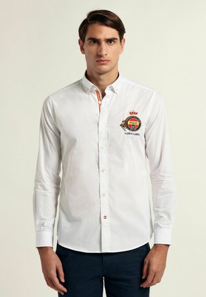 Camisa blanca de manga larga con cuello abotonado, un detalle de rayas de colores y un logo bordado en el pecho. Con cuello y cierre de botón.