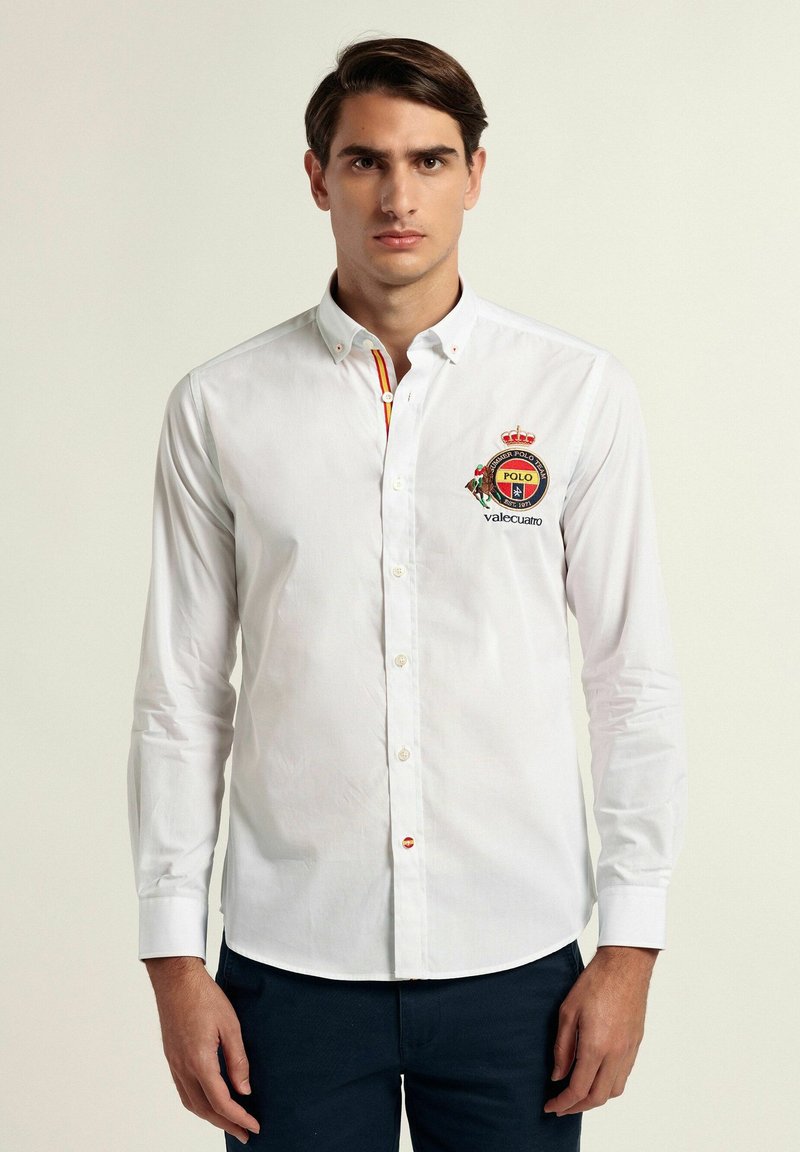 Camisa blanca de manga larga con cuello abotonado, un detalle de rayas de colores y un logo bordado en el pecho. Con cuello y cierre de botón.