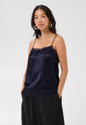 Femme portant un caraco en satin bleu marine avec bordure en dentelle et pantalon noir, debout les mains dans les poches devant un fond uni.