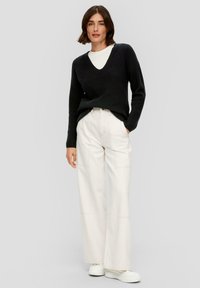 Schwarzer Strick-V-Ausschnitt-Pullover kombiniert mit cremefarbenen, weiten Hosen. Das Model trägt weiße Plateausneaker. Einfaches und sauberes Design, ohne Muster.