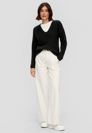 Schwarzer Strick-V-Ausschnitt-Pullover kombiniert mit cremefarbenen, weiten Hosen. Das Model trägt weiße Plateausneaker. Einfaches und sauberes Design, ohne Muster.
