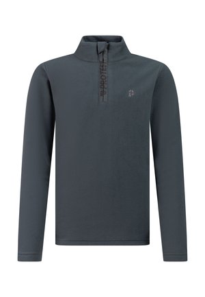 Jong persoon in een zwarte muts, grijs langärmelig vest met rits, zwarte broek en zwarte schoenen staand met de handen in de zij, kijkt zijwaarts.