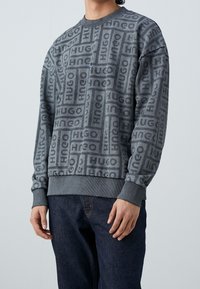 Sudadera gris con un patrón repetido de logotipos "HUGO", puños y dobladillo de canalé, y un cuello redondo. Combinada con jeans oscuros.