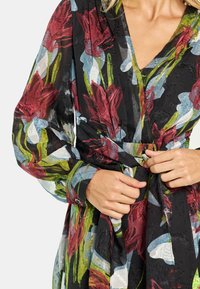 Vestido envuelto floral hecho de tela negra translúcida con patrones de flores en rojo, azul y verde. Incluye un cinturón de lazo y mangas largas y voluminosas.